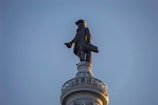 A statue of a historical figure in traditional attire stands atop a column-like structure against a clear sky. The figure is wearing a hat and carrying an object under one arm.