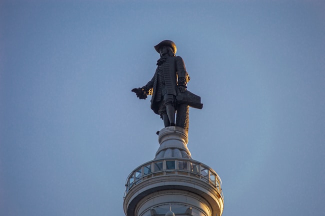 A statue of a historical figure in traditional attire stands atop a column-like structure against a clear sky. The figure is wearing a hat and carrying an object under one arm.
