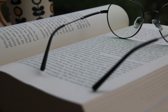 An open book with pages filled with text, accompanied by a pair of round eyeglasses resting on top. In the background, there is a blurred mug with a flower pattern, suggesting a relaxed reading environment.