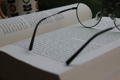 An open book with pages filled with text, accompanied by a pair of round eyeglasses resting on top. In the background, there is a blurred mug with a flower pattern, suggesting a relaxed reading environment.