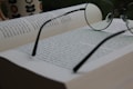An open book with pages filled with text, accompanied by a pair of round eyeglasses resting on top. In the background, there is a blurred mug with a flower pattern, suggesting a relaxed reading environment.
