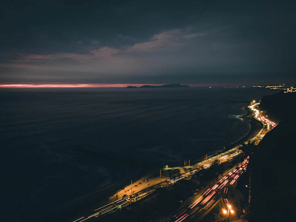 The image depicts a coastal city at night, with a dark sky and silhouetted buildings on the left side. The cityscape is illuminated by streetlights and car headlights, creating a contrast between the natural darkness of the night sky and the artificial brightness of the urban environment. In the distance, the ocean stretches out to meet the horizon under a cloudy sky, adding depth and perspective to the scene.