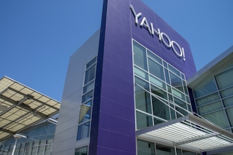 A modern building with a prominent purple panel displaying the word 'YAHOO!' in large letters. The structure features large glass windows and a metal awning, set against a clear blue sky.