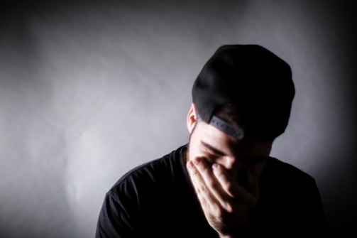 A person wearing a black cap and black shirt is covering their face with one hand. The lighting creates a somber atmosphere, highlighting the emotional expression.