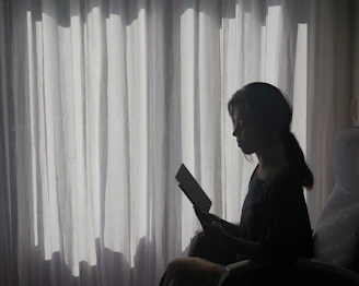 A shadowy silhouette of a woman reading by moonlight, framed by sheer curtains.