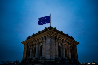 A dark structure topped with a European Union flag flying against a dimly lit sky. The building has columns and an ancient architectural style, suggesting a historical or governmental significance.