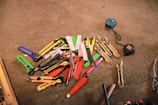 A collection of various utility knives and box cutters in different colors, such as red, yellow, green, and blue, placed on a concrete surface. Two measuring tapes are positioned to the side, while a pair of pliers is visible near the bottom edge.