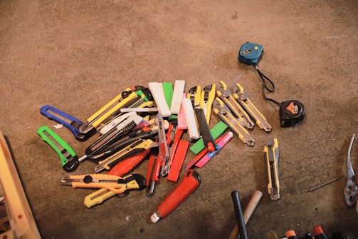 A collection of various utility knives and box cutters in different colors, such as red, yellow, green, and blue, placed on a concrete surface. Two measuring tapes are positioned to the side, while a pair of pliers is visible near the bottom edge.