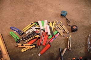 A collection of various utility knives and box cutters in different colors, such as red, yellow, green, and blue, placed on a concrete surface. Two measuring tapes are positioned to the side, while a pair of pliers is visible near the bottom edge.