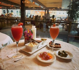 A table set with a variety of appetizers and two vibrant red cocktails garnished with fruit, placed on an elegantly embroidered tablecloth. Small plates feature grapes, cheese, seasoned olives, chocolates, and pastries. The table is adorned with a small bouquet of flowers and a candle. In the background, there is a view into a busy restaurant with numerous diners visible through large windows.