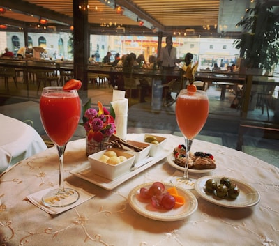 A table set with a variety of appetizers and two vibrant red cocktails garnished with fruit, placed on an elegantly embroidered tablecloth. Small plates feature grapes, cheese, seasoned olives, chocolates, and pastries. The table is adorned with a small bouquet of flowers and a candle. In the background, there is a view into a busy restaurant with numerous diners visible through large windows.