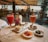 A table set with a variety of appetizers and two vibrant red cocktails garnished with fruit, placed on an elegantly embroidered tablecloth. Small plates feature grapes, cheese, seasoned olives, chocolates, and pastries. The table is adorned with a small bouquet of flowers and a candle. In the background, there is a view into a busy restaurant with numerous diners visible through large windows.