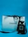 A black Chanel perfume bottle is placed next to a white Chanel shopping bag with black handles. The background is a solid blue color, creating a contrast that highlights the elegance of the items.