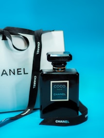 A black Chanel perfume bottle labeled 'Coco Noir' stands in front of a white Chanel shopping bag with black handles. A black ribbon with Chanel branding loops around both the bottle and bag against a bright blue background.