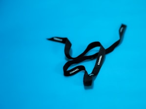 A black ribbon with 'CHANEL' written in white letters against a bright blue background. The ribbon appears loosely arranged and has a luxurious texture.