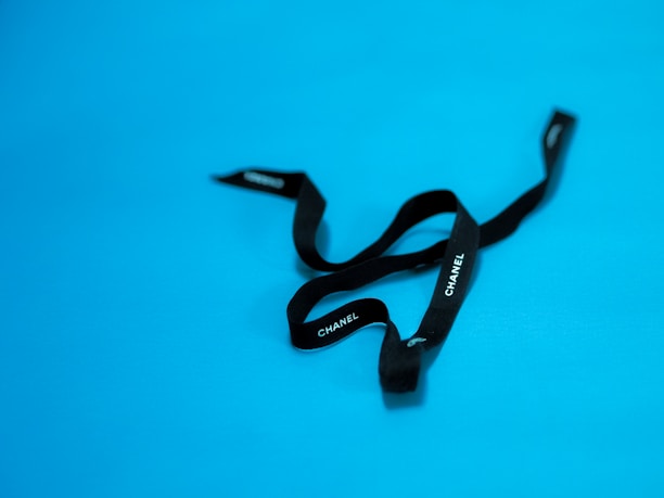 A black ribbon with 'CHANEL' written in white letters against a bright blue background. The ribbon appears loosely arranged and has a luxurious texture.