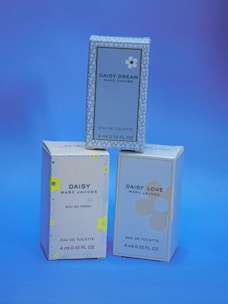 Three small boxes of Marc Jacobs fragrances are displayed against a blue background. The boxes are labeled 'Daisy Dream,' 'Daisy,' and 'Daisy Love,' each with corresponding floral designs.