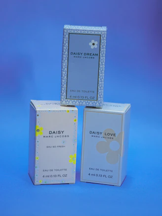 Three small boxes of Marc Jacobs fragrances are displayed against a blue background. The boxes are labeled 'Daisy Dream,' 'Daisy,' and 'Daisy Love,' each with corresponding floral designs.