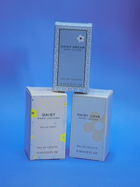 Three small boxes of Marc Jacobs fragrances are displayed against a blue background. The boxes are labeled 'Daisy Dream,' 'Daisy,' and 'Daisy Love,' each with corresponding floral designs.