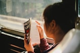 A person is reading a book while sitting next to a window. The book cover displays the title 'The Girl Who Played with Fire' by Stieg Larsson. The surroundings outside the window appear to be blurred.