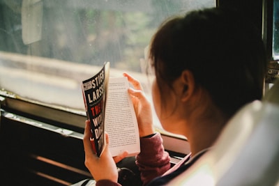 A person is reading a book while sitting next to a window. The book cover displays the title 'The Girl Who Played with Fire' by Stieg Larsson. The surroundings outside the window appear to be blurred.