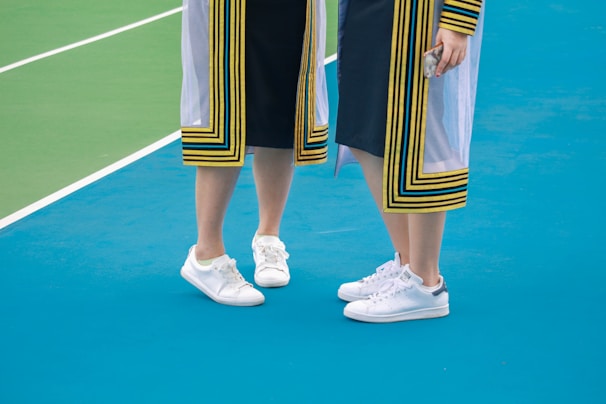 Two people standing on a vibrant blue surface, wearing academic gowns with yellow and black stripes on the edges. They are both wearing white sneakers, and one is holding a phone in their right hand.