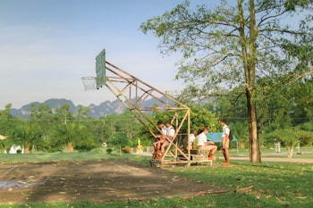 A group of school-aged boys in uniforms are sitting and standing around a simple, makeshift bench under a metal basketball hoop. The scene is set outdoors in a grassy area with trees and mountains visible in the background. The overall environment is lush and peaceful, with the afternoon sunlight creating long shadows.