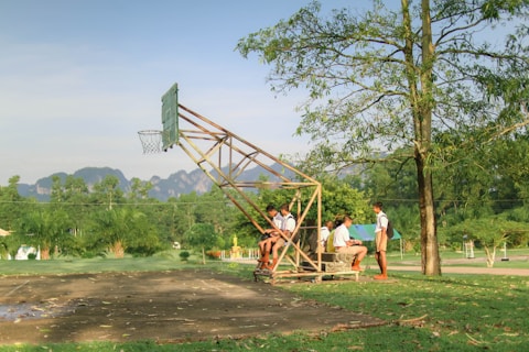 A group of school-aged boys in uniforms are sitting and standing around a simple, makeshift bench under a metal basketball hoop. The scene is set outdoors in a grassy area with trees and mountains visible in the background. The overall environment is lush and peaceful, with the afternoon sunlight creating long shadows.