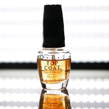 A clear bottle of OPI top coat nail polish stands on a reflective surface. The label on the bottle is visible with black text, and there is a dark-colored cap on top. The background is bright and evenly lit, enhancing the visibility of the product.