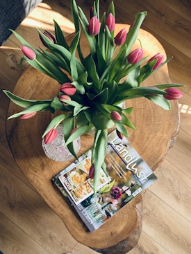 A wooden table holds a vase of pink tulips with lush green leaves. Beside the vase, there is a magazine titled 'Landlust' featuring a cover image of baked goods and plants.