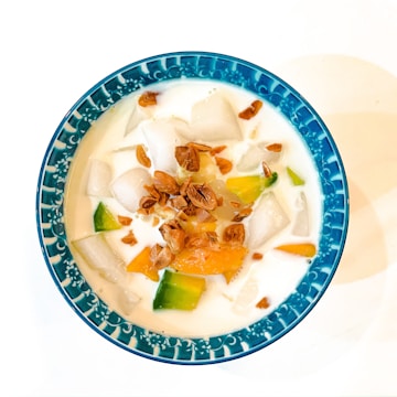 A colorful dessert in a blue ceramic bowl, featuring a mixture of white milky liquid, white jelly-like cubes, orange and green jelly pieces, topped with small brown caramelized coconut bits.