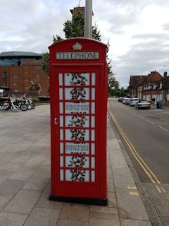 A traditional red telephone booth is repurposed with signage for the Stratford Coffee Box. The booth features images of coffee beans on the glass panes. It is situated on a paved pedestrian area with a background of motorcycles, buildings, and trees.