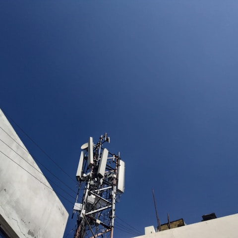 A telecommunications tower equipped with various antennas and equipment surrounded by buildings. The sky is clear and blue, providing a stark, expansive background.