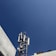 A telecommunications tower equipped with various antennas and equipment surrounded by buildings. The sky is clear and blue, providing a stark, expansive background.
