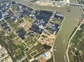 An aerial view of agricultural fields with a river cutting through the landscape. The fields are divided into rectangular patches, some covered with dark-colored materials, possibly solar panels or greenhouses. Several boats are visible on the river, suggesting transportation or trade activity. Infrastructure like roads and buildings are also visible, indicating human settlement and development.