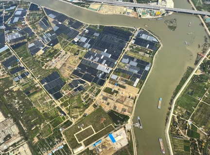 An aerial view of agricultural fields with a river cutting through the landscape. The fields are divided into rectangular patches, some covered with dark-colored materials, possibly solar panels or greenhouses. Several boats are visible on the river, suggesting transportation or trade activity. Infrastructure like roads and buildings are also visible, indicating human settlement and development.