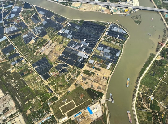 An aerial view of agricultural fields with a river cutting through the landscape. The fields are divided into rectangular patches, some covered with dark-colored materials, possibly solar panels or greenhouses. Several boats are visible on the river, suggesting transportation or trade activity. Infrastructure like roads and buildings are also visible, indicating human settlement and development.