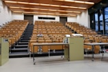 A large, empty lecture hall with tiered wooden seating and a single person sitting in the middle row. The room features a beige color scheme with overhead lights and large windows on the right side, allowing natural light. Two podiums and desks with electronic equipment are positioned at the front.