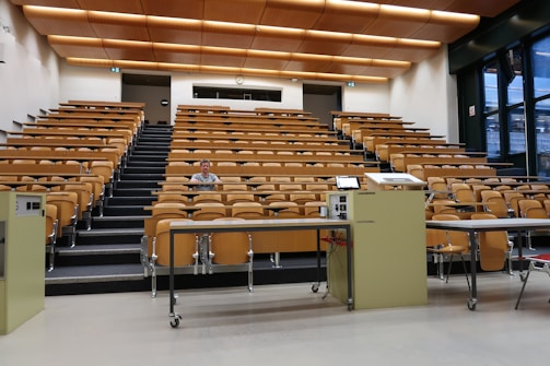 A large, empty lecture hall with tiered wooden seating and a single person sitting in the middle row. The room features a beige color scheme with overhead lights and large windows on the right side, allowing natural light. Two podiums and desks with electronic equipment are positioned at the front.