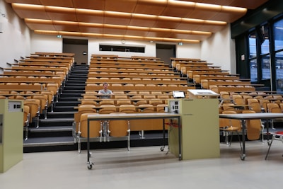 A large, empty lecture hall with tiered wooden seating and a single person sitting in the middle row. The room features a beige color scheme with overhead lights and large windows on the right side, allowing natural light. Two podiums and desks with electronic equipment are positioned at the front.