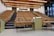 A large, empty lecture hall with tiered wooden seating and a single person sitting in the middle row. The room features a beige color scheme with overhead lights and large windows on the right side, allowing natural light. Two podiums and desks with electronic equipment are positioned at the front.