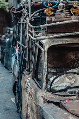An old, rusted vehicle covered in various items and adorned with gold-colored flowers and a small statue on its roof. The vehicle appears weathered and abandoned, with ropes and other paraphernalia attached to it.