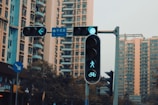 Urban setting with a focus on a traffic light showing green signals for pedestrians and cyclists. High-rise buildings form the backdrop along with some street signs.