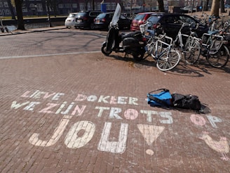 Chalk writing on a brick pavement reads 'Lieve dokter, we zijn trots op jou!' with a drawing of a chicken and an exclamation mark. A scooter is parked on the pavement alongside several bicycles. A backpack is lying on the ground, and cars are visible in the background under sunny weather.