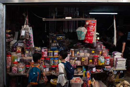 A small shop packed with various goods, including jars of candies, snacks, and traditional items. Hanging above are decorative items and lanterns. Two children with backpacks stand in front, observing the assortment. The shop interior is dimly lit, adding a nostalgic and cozy vibe.
