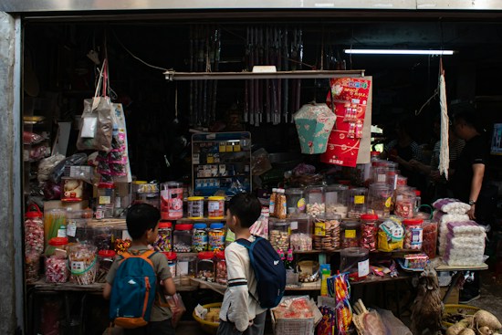 A small shop packed with various goods, including jars of candies, snacks, and traditional items. Hanging above are decorative items and lanterns. Two children with backpacks stand in front, observing the assortment. The shop interior is dimly lit, adding a nostalgic and cozy vibe.