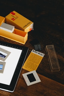 A wooden table holds a yellow Kodak color slides box, an open compartment with additional slides, a graphic tablet, slides with images, and a yellow paper labeled 'Need Help?'.