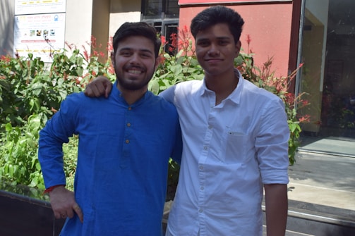 Two young men are standing closely with their arms around each other, smiling at the camera. They are in a garden setting with green foliage and red flowers in the background. One man is wearing a blue shirt while the other is wearing a white shirt.