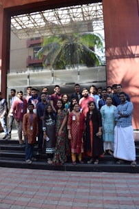A group of people are standing on steps in front of a large glass-fronted building. They are wearing a mix of traditional and modern attire, suggesting a diverse and multicultural gathering. Behind them, a palm tree is visible through the glass, reflecting the outdoor environment.