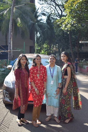 Four women are standing together in an outdoor setting, with trees and a building in the background. They are wearing colorful traditional attire and appear to be smiling. A car is parked behind them, partially visible.